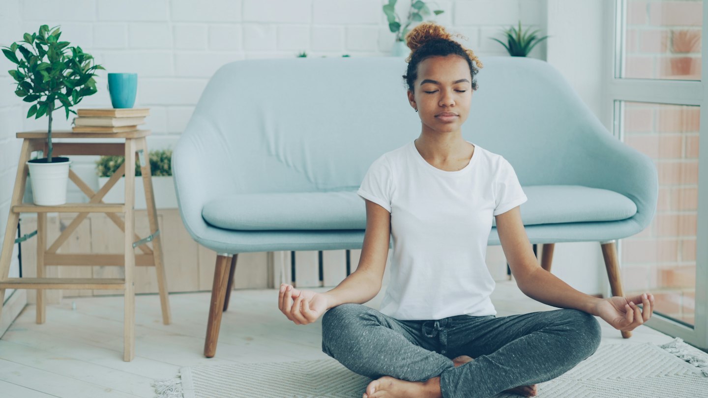 Donna seduta in posizione del loto su un tappeto in un soggiorno, in meditazione.